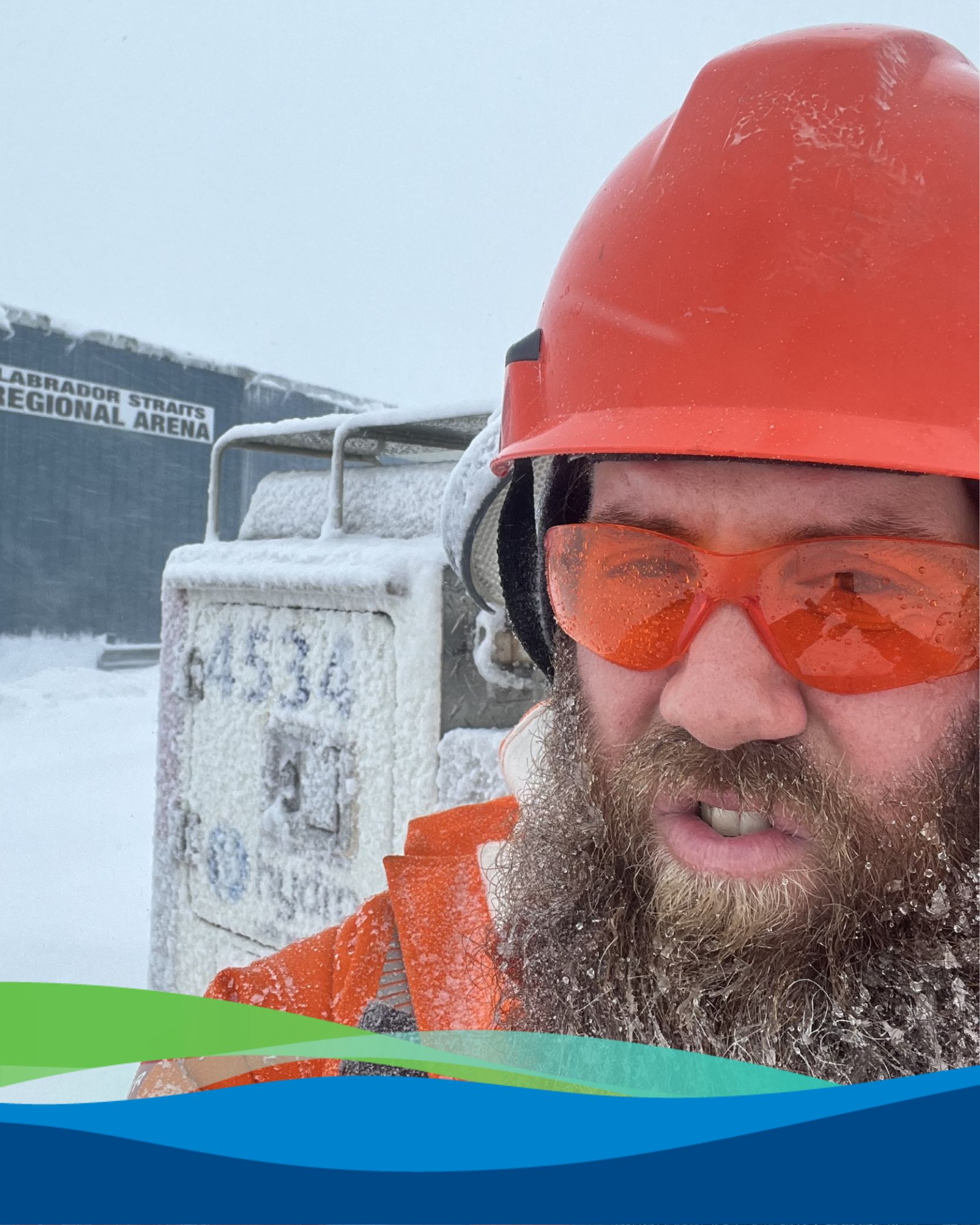 Damien is wearing an orange hard hat, safety glasses and winter jacket. He has snow in his beard and is standing in front of a snow covered Hydro vehicle near the Labrador Straits Regional Arena