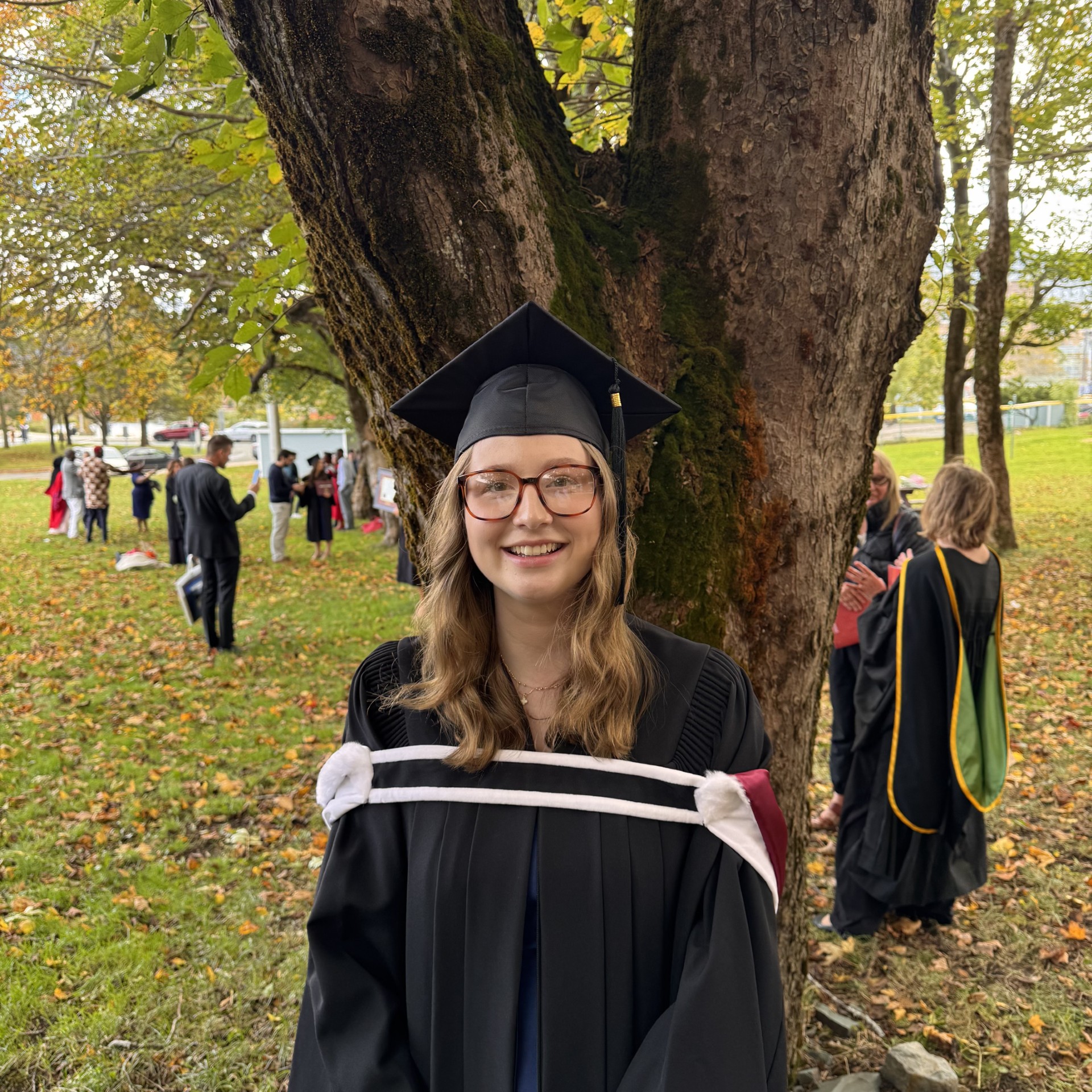 Jadzia is wearing glasses and smiling at the camera as she wears her graduation cap and gown, standing outside in front of a tree