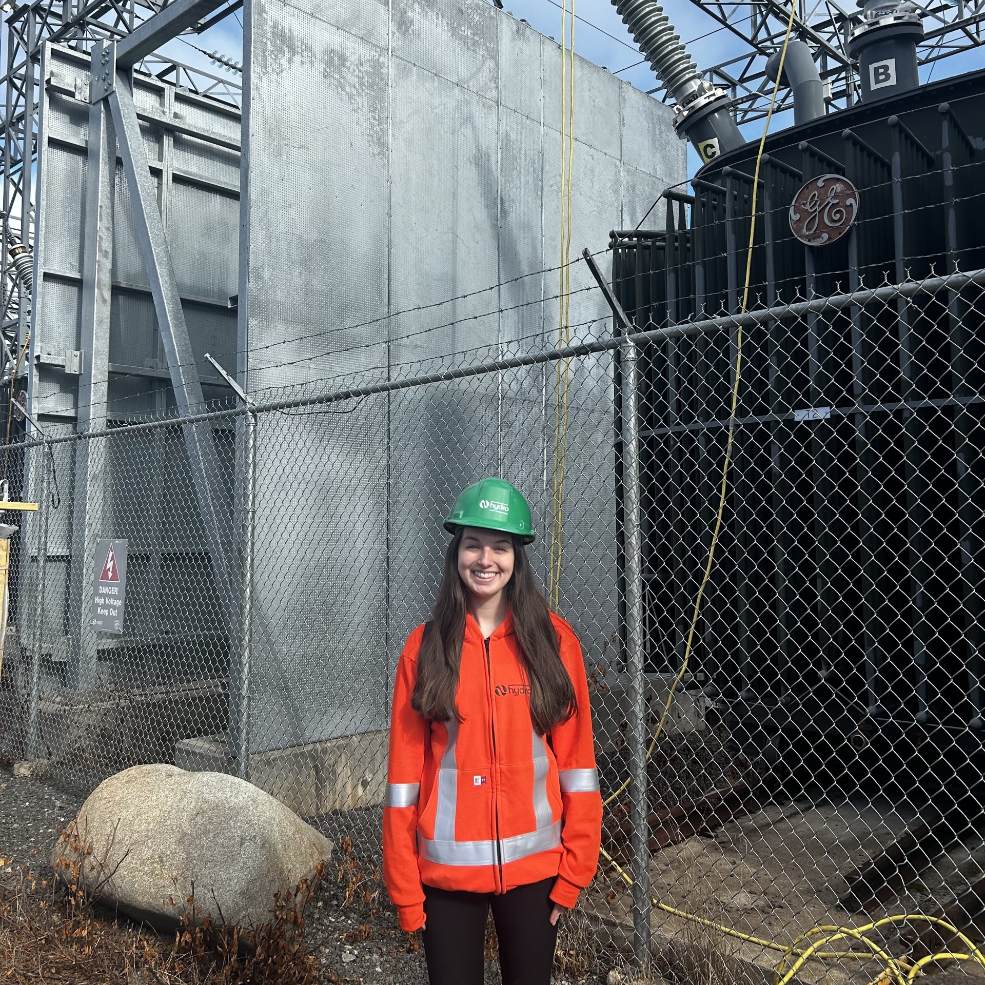 Jayda is wearing PPE and standing in front of a grey concrete wall and chain link fence