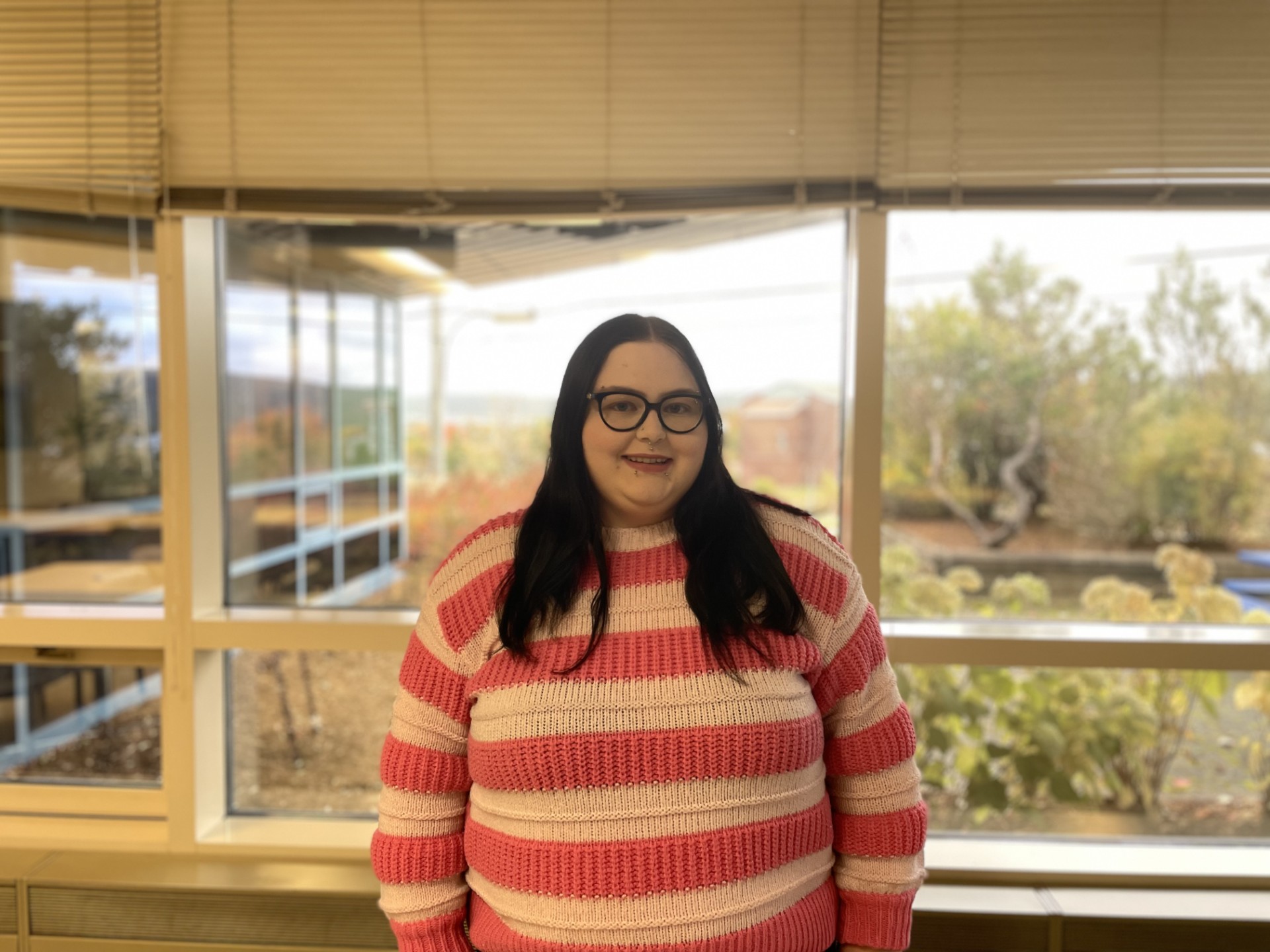Logan is standing in front of a bright window, wearing glasses and a striped pink sweater