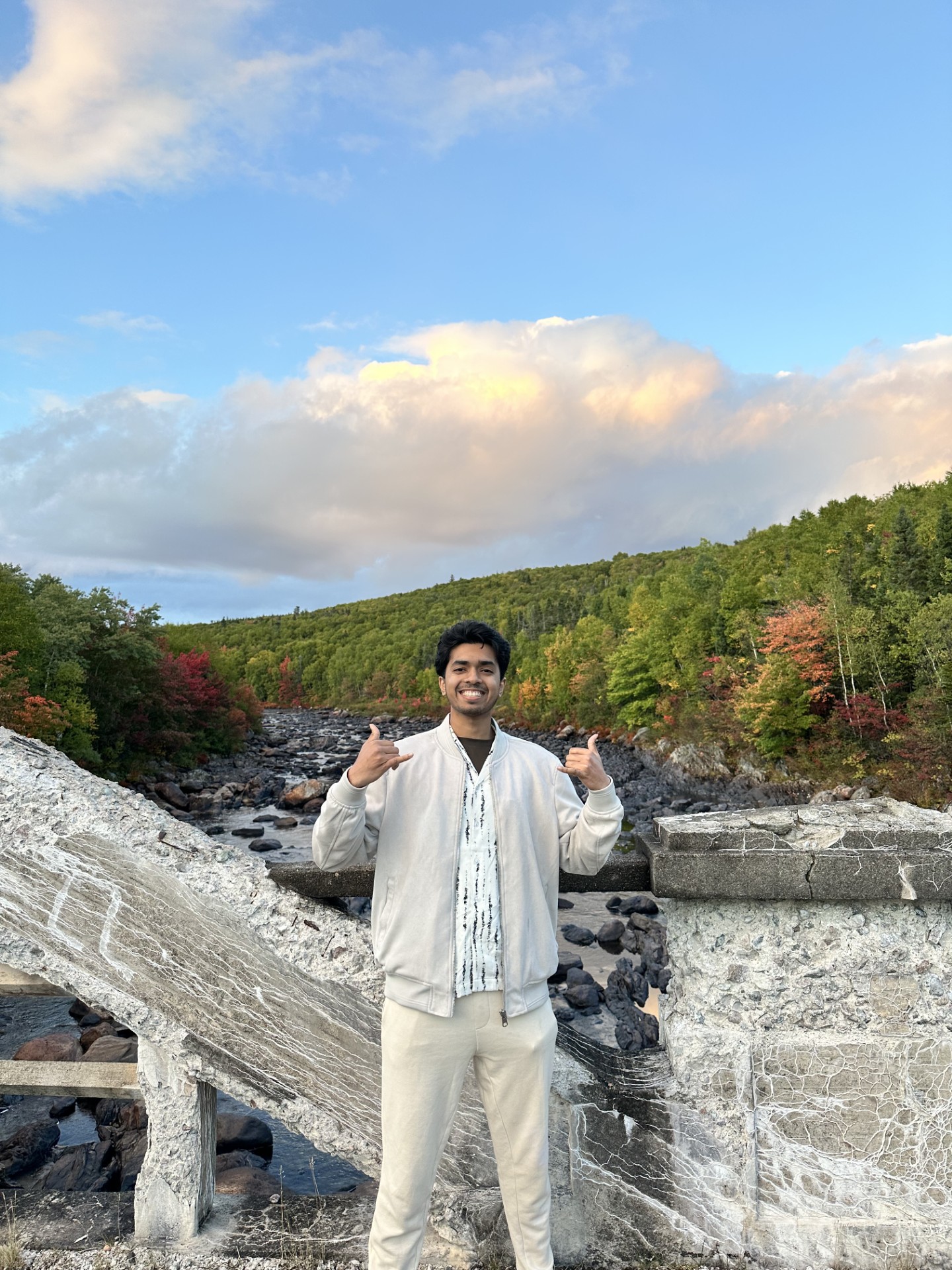 Taj stands in front of a lovely natural landscape with trees and a river, as he gives a double thumbs up to the camera