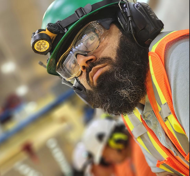 An artsy shot shows a close up of Tarik in full PPE in a work environment as he looks away from the camera
