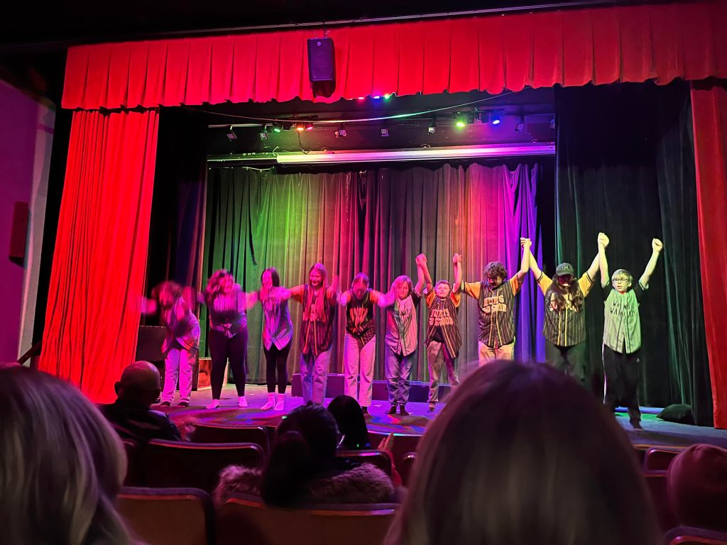 A group of performers stands on stage, holding hands and bowing to the audience. The backdrop features colorful stage lights in red, green, and purple, while seated audience members are partially visible in the foreground.