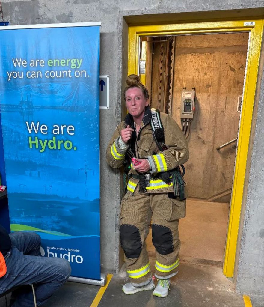 A stair climb participant leans against a doorway following the climb. She is wearing fire fighting gear, but no helmet. Next to her is a banner that reads We are energy you can count on. We are Hydro