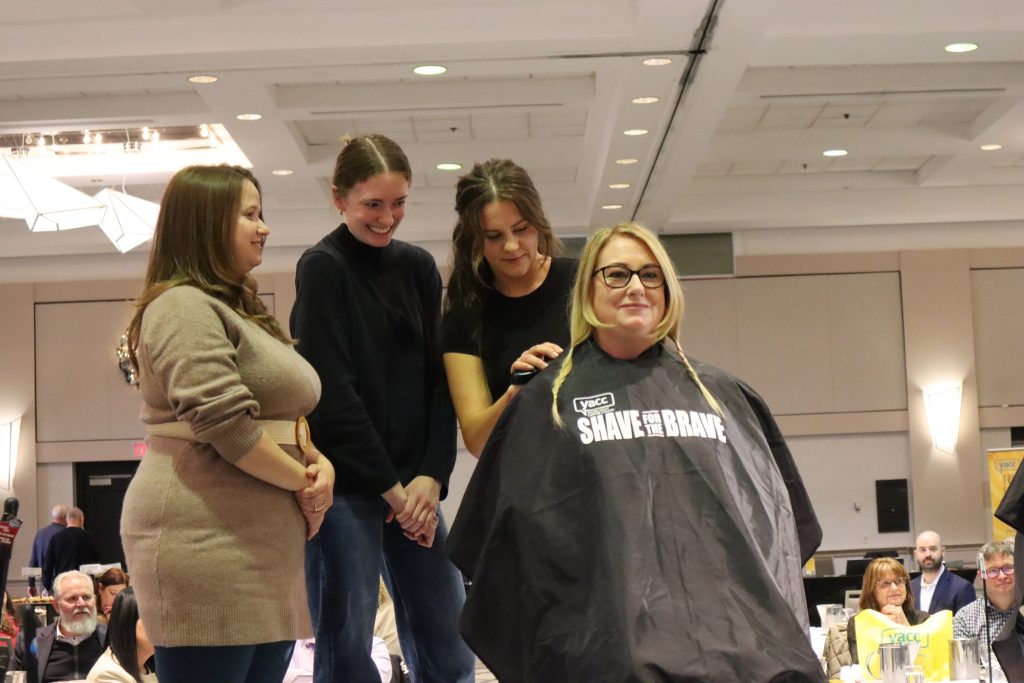 Jennifer Williams sits in the foreground wearing a grey hair dresser cape. Her hair is in several braids. Behind her a hair dresser cuts a braid while two women look on.