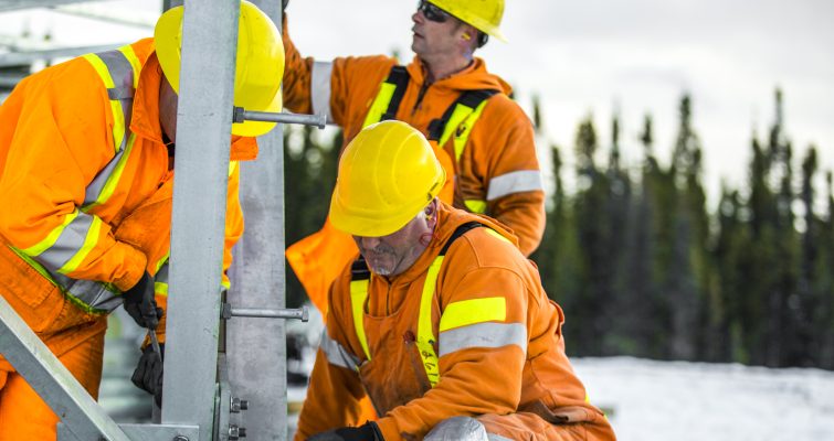 Three workers in yellow hard hats and orange PPE working on the base of a transmission tower.