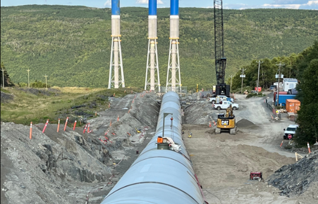 A long penstock pipe with gravel on either side. Three stacks can be seen in the background.