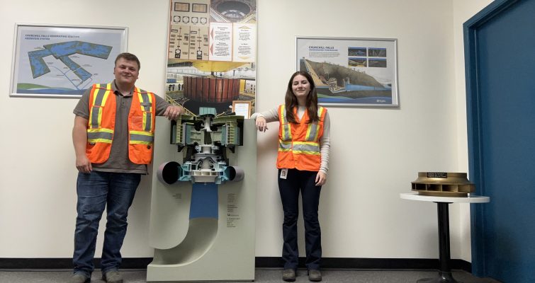 Two Hydro employees in safety vests smiling at the camera while standing in an office next to an informational banner about a piece of equipment.