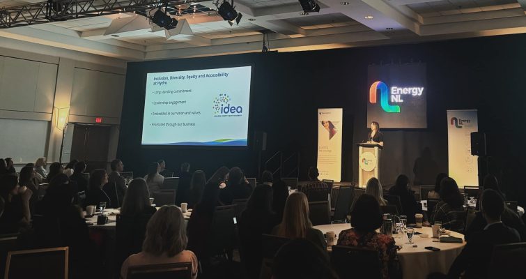 Amanda McClafferty speaking on stage at the event. An Energy NL logo is behind her and a slide presentation is displayed beside her. Audience member's heads can be seen in the foreground.