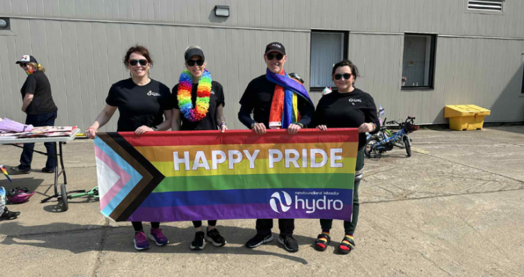 Churchill Falls employees stand outside a building holding a rainbow banner that says Happy Pride and features the Hydro logo.