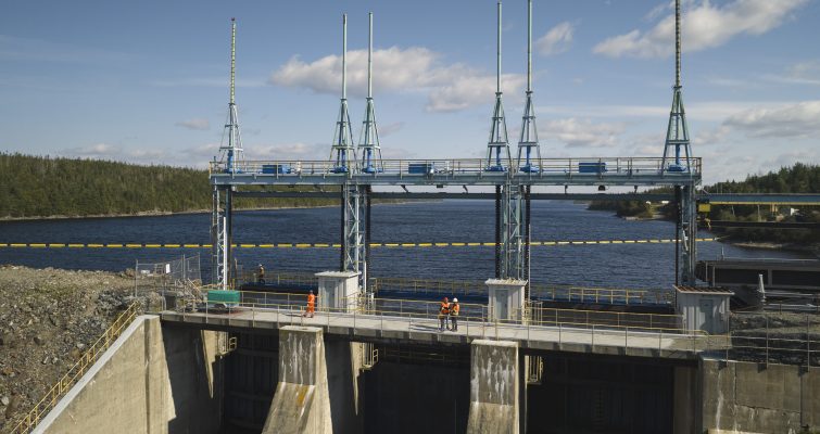 Large concrete dam at Hydro's Bay d'Espoir facility. Four employees in orange PPE and hard hats are standing on top. Green hills, blue sky and clouds in the background.