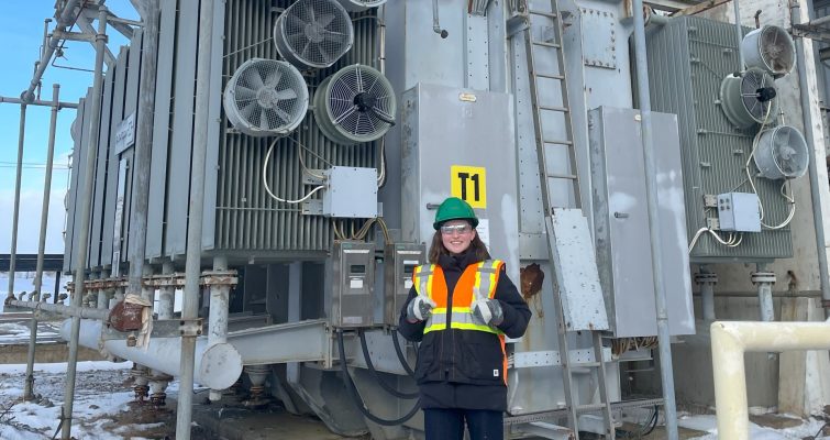 Student in green hard hat and safety vest standing in front of electrical equipment in a switchyard. There is snow on the ground.
