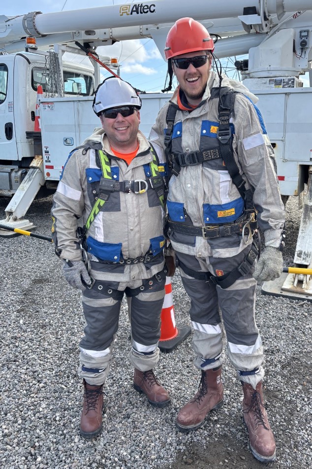 Two powerline technicians dressed in safety gear with hard hats and fall arrest harnesses stand in front of a boom truck.