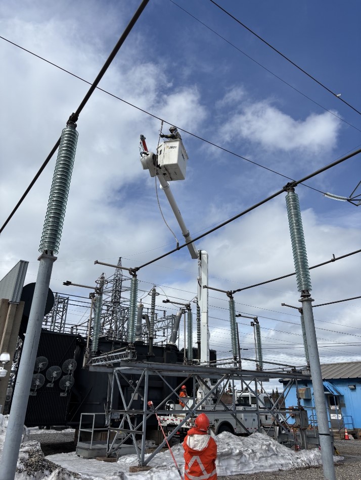 A powerline technician performs live line work in an electrical yard from a boom truck. Below another powerline technician looks on.