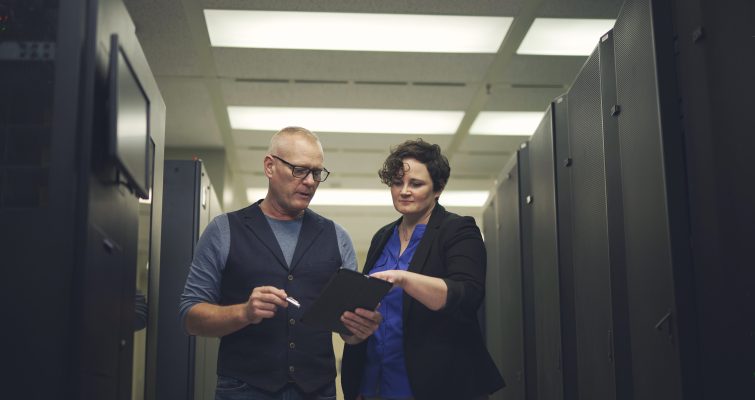 Two employees looking and pointing at an iPad. Computer servers can be seen behind them.