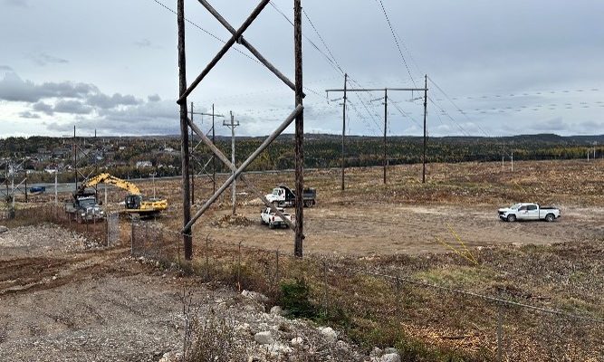 Transmission line construction area with utility vehicles, an excavator, and power infrastructure in a cleared field beneath a cloudy sky.